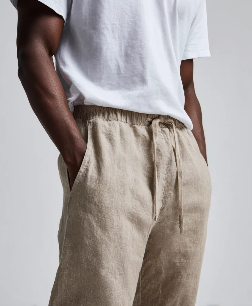 Close-up of a man's torso, showcasing beige linen drawstring pants and a plain white t-shirt.  His hands are casually in his pockets. The image highlights the relaxed fit and texture of the linen pants. The overall style is simple and casual.