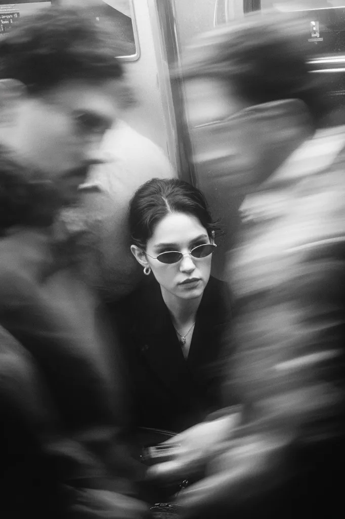 A young woman with dark hair and oval sunglasses sits on a public transport, her expression neutral.  Surrounding her, blurred figures suggest the rush and movement of commuters. The monochrome image creates a sense of anonymity and fleeting moments within a busy urban setting. The contrast between the sharp focus on the woman and the blurred background emphasizes her stillness amidst the chaos.