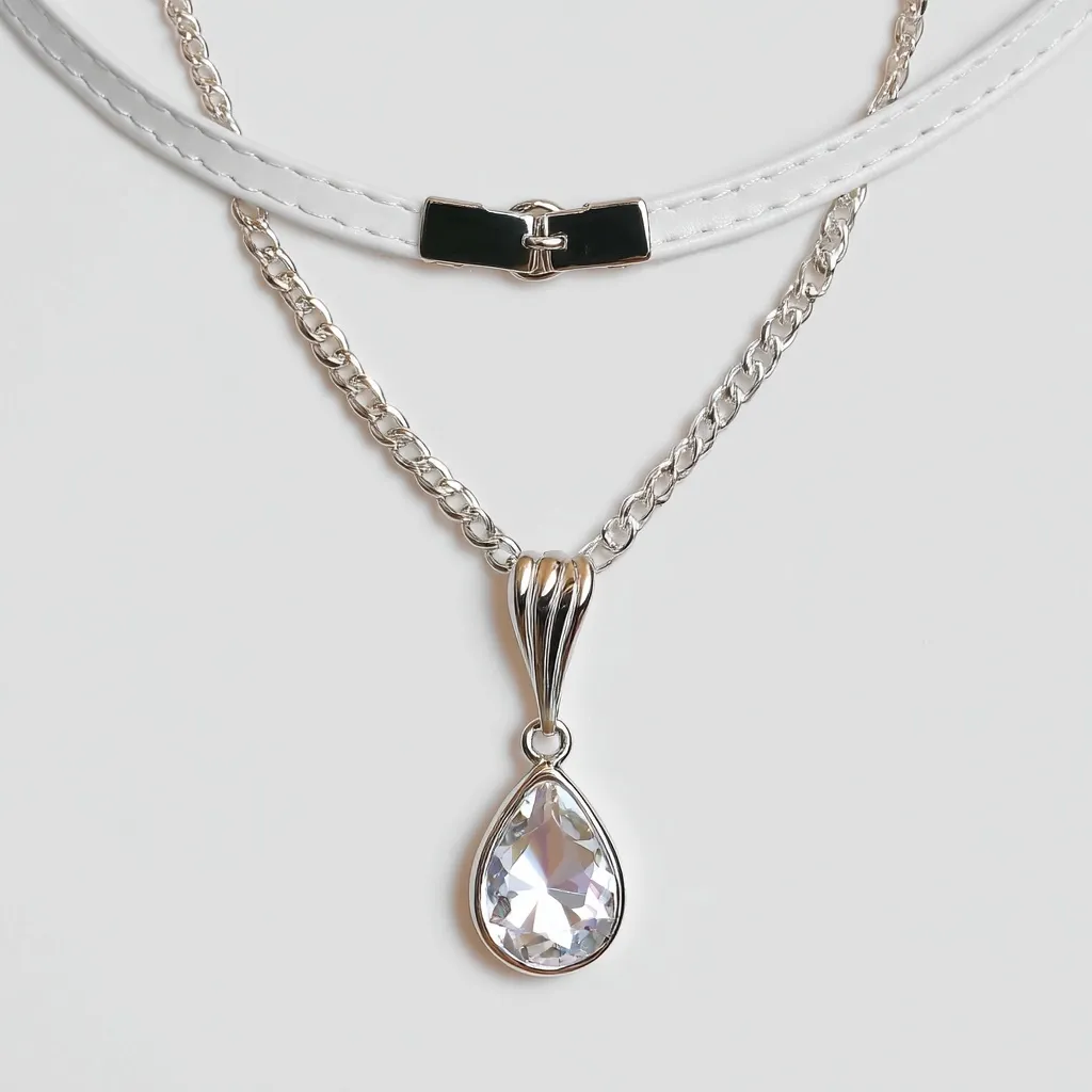 An elegant layered necklace features a white leather choker with a black clasp, and a delicate silver chain adorned with a pear-shaped clear crystal pendant. The pendant hangs gracefully from the chain, complementing the simple yet stylish design. The necklace is displayed against a white backdrop, highlighting its minimalist aesthetic.  The overall effect is one of refined sophistication.
