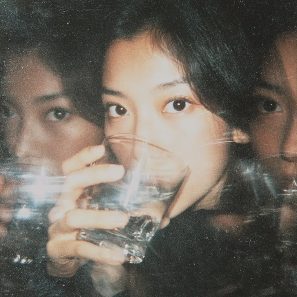 Close-up shot of a young woman with dark hair, holding a glass of liquid to her lips.  The image has a grainy, vintage filter applied, creating a nostalgic aesthetic.  Partial reflections of other faces are visible, adding to the blurry, dreamlike quality. The overall mood is intimate and mysterious.