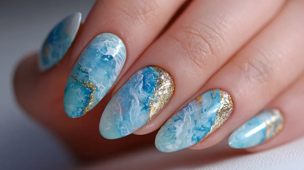 Close-up of elegantly manicured nails.  Each nail features a unique design of light blue and white marbling, accented with gold foil detailing, creating a beautiful ocean-inspired effect.  The almond-shaped nails are long and perfectly polished, showcasing a sophisticated and trendy nail art style.
