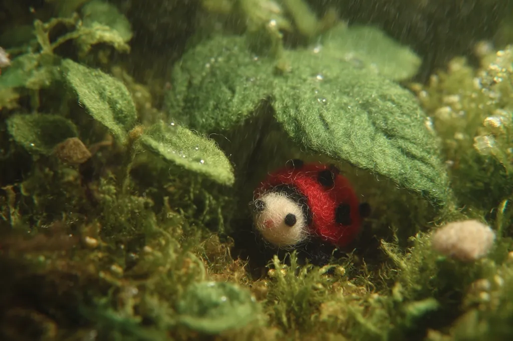 A miniature needle-felted ladybug, with a white face and red shell, seeks shelter under a large green leaf in a lush, mossy environment.  Rain gently falls, creating a serene and whimsical scene. The vibrant green foliage contrasts beautifully with the ladybug's bright colors. The overall image evokes a feeling of tranquility and the beauty of nature in miniature.