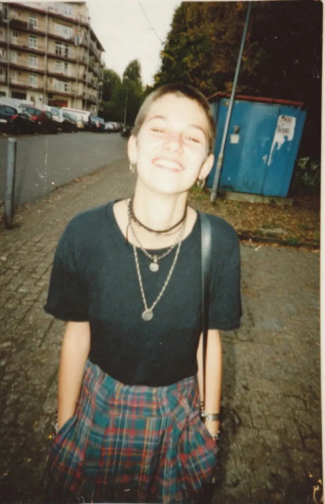 A young woman with short, buzzed hair smiles for the camera. She wears a black t-shirt, a red, green, and blue plaid mini-skirt, and several necklaces.  A black shoulder bag hangs from her shoulder. The photo appears to be taken outdoors, near a street with parked cars and a building in the background. The image has a vintage, slightly grainy quality suggesting it's an older photograph.