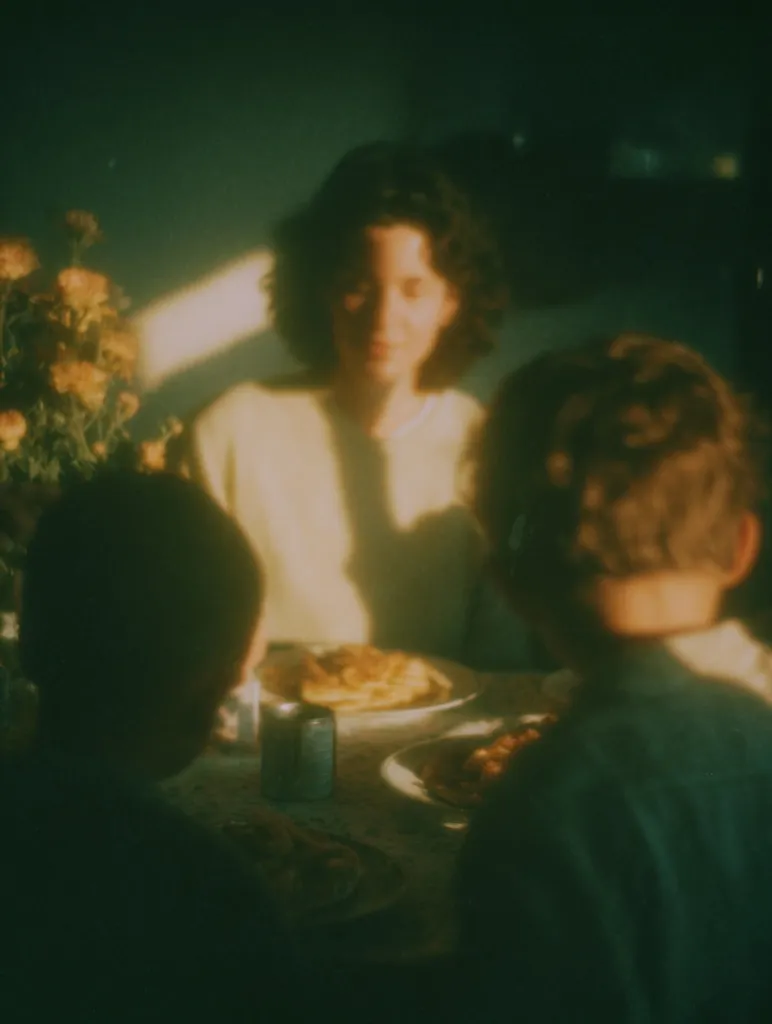 A sun-drenched photograph captures a family meal. A woman with dark hair sits at a table, bathed in warm light, with two children seated beside her.  The scene is blurry, adding to its nostalgic and intimate feel. Plates of food are visible on the table, and a bouquet of flowers sits nearby. The overall impression is one of quiet domesticity and golden hour warmth.