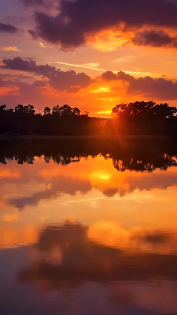 The setting sun casts a golden glow on the lake, reflecting off the surface of the water and illuminating the trees on the shore. The sky is ablaze with color, the most vibrant oranges and yellows imaginable. The scene is one of peace and tranquility, and the viewer is invited to relax and enjoy the beauty of nature.